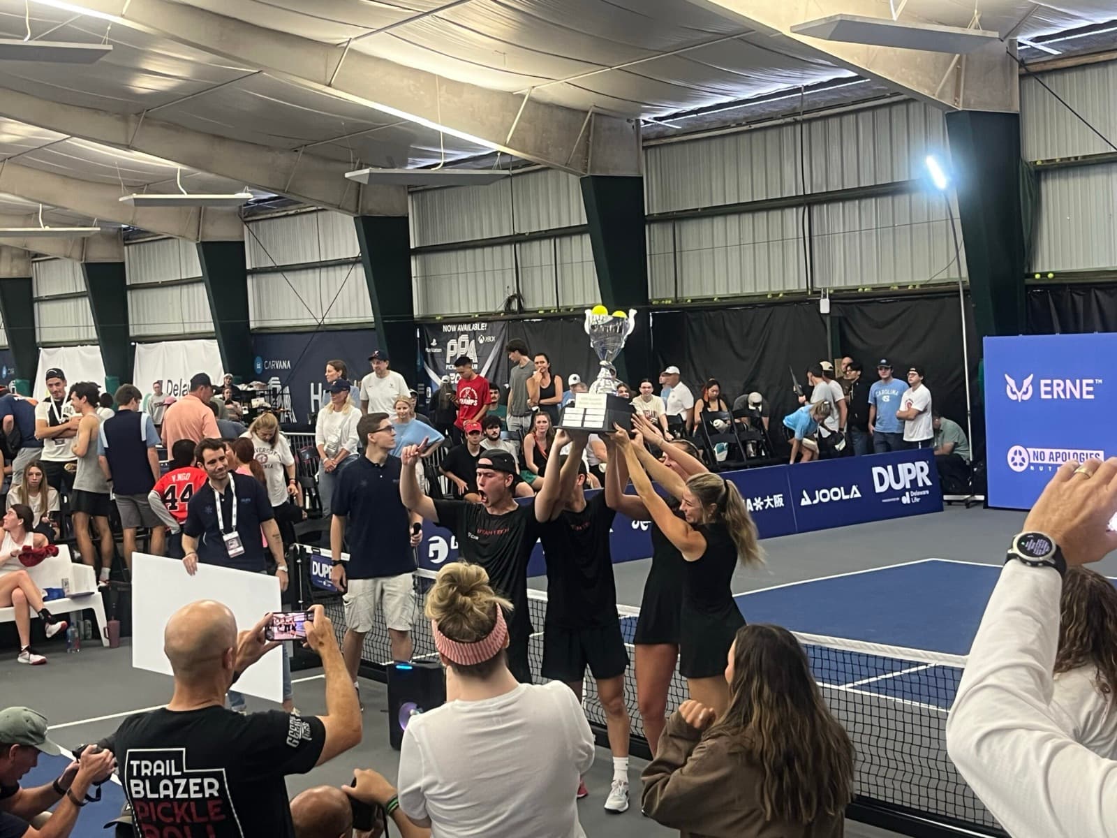 A collegiate doubles team raises a championship trophy overhead, gold medals around their necks.