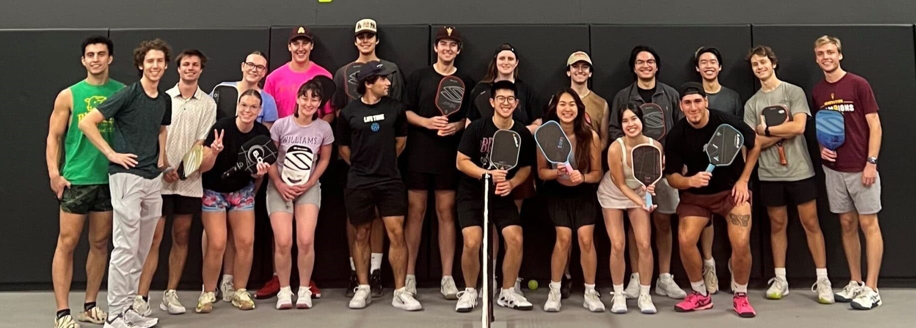 A collegiate pickleball club lines up together indoors for a group photo, paddles in hand.